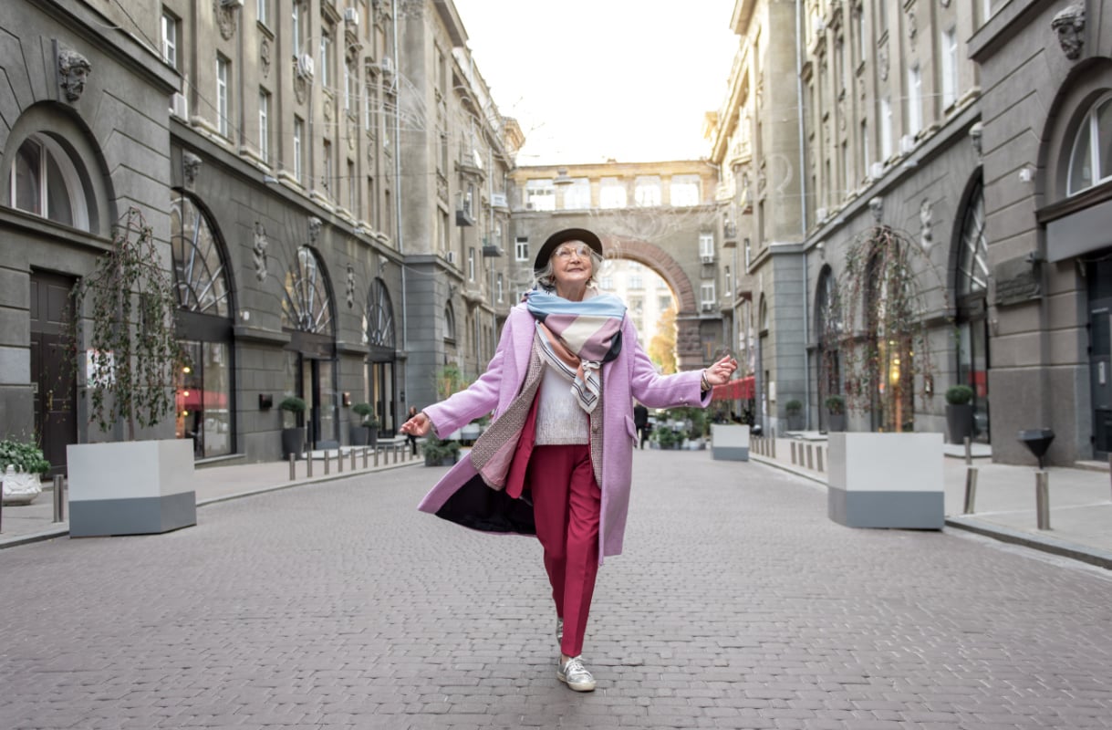 Iäkäs nainen värikkäissä vaatteissa kävelemässä iloisena kaupungilla.