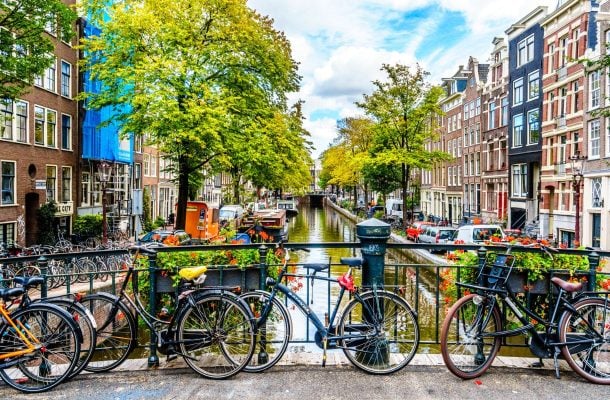 Useita polkupyöriä nojailemassa kanaalin ylittävään sillan kaiteeseen Amsterdamissa. Taustalla kanaali, jonka molemmin puolin on värikkäitä kerrostaloja ja vehreitä puita.