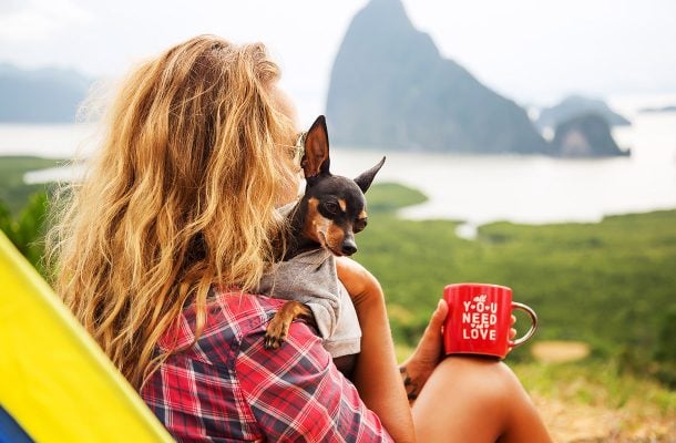Nainen istumassa teltan edessä koira sylissä ja kahvikuppi kädessä. Taustalla kohoaa vuori. 