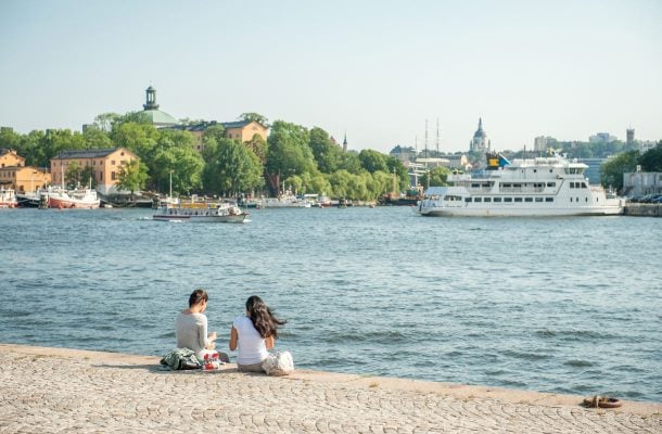 Kaksi ihmistä istumassa laiturilla saariston äärellä Tukholmassa, taustalla saari ja laiva.